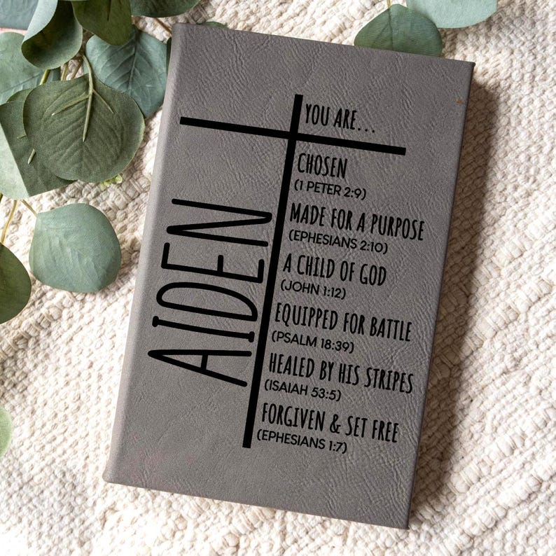 May include: Gray journal with the name "AIDEN" vertically on the left side. The journal features a cross design and inspirational text including "YOU ARE... CHOSEN" and "MADE FOR A PURPOSE". The journal is on a white textured surface.