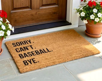 Może przedstawiać: Prostokątna wycieraczka z włókna kokosowego z czarnym napisem "SORRY. CAN'T. BASEBALL. BYE.". Mata leży na szarej kamiennej powierzchni w pobliżu drewnianych drzwi. Widoczne są rośliny doniczkowe.