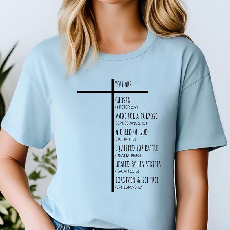 May include: A light blue t-shirt with a black cross and text that reads "You are... Chosen (1 Peter 2:9) Made for a Purpose (Ephesians 2:10) A Child of God (John 1:12) Equipped for Battle (Psalm 18:39) Healed by His Stripes (Isaiah 53:5) Forgiven & Set Free (Ephesians 1:7)"