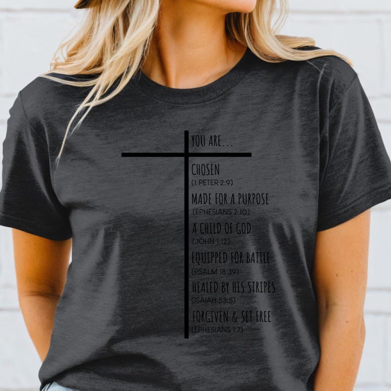 May include: A dark gray t-shirt with a black cross and text that reads "You are... Chosen (1 Peter 2:9) Made for a Purpose (Ephesians 2:10) A Child of God (John 1:12) Equipped for Battle (Psalm 18:39) Healed by His Stripes (Isaiah 53:5) Forgiven & Set Free (Ephesians 1:7)"