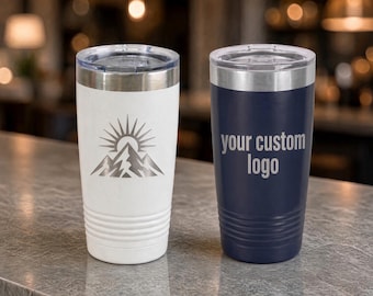 May include: Two stainless steel tumblers with clear plastic lids and ribbed bases. The white tumbler features a mountain and sun graphic, while the navy blue tumbler displays the text your custom logo engraved on the front surface.