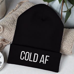 Może przedstawiać: Czarna czapka typu beanie z białym napisem "COLD AF". Czapka jest złożona na brzegu i wykonana z miękkiego, dzianinowego materiału. Czapka leży na beżowym kocu.