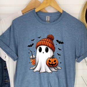 May include: A blue t-shirt featuring a cartoon ghost wearing an orange and white knitted hat and holding a pumpkin and a cup of orange juice. The ghost is surrounded by bats and stars.
