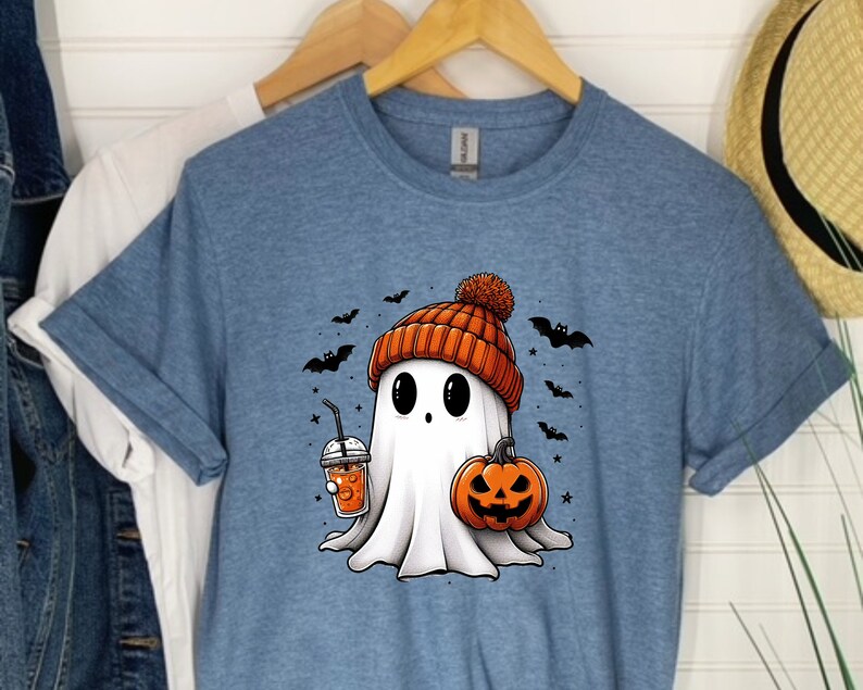 May include: A blue t-shirt featuring a cartoon ghost wearing an orange and white knitted hat and holding a pumpkin and a cup of orange juice. The ghost is surrounded by bats and stars.