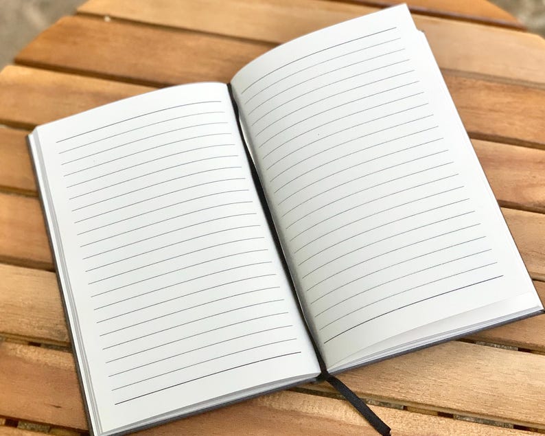 May include: A black lined notebook with a white cover and a black spine. The notebook is open to a blank page with lined paper.