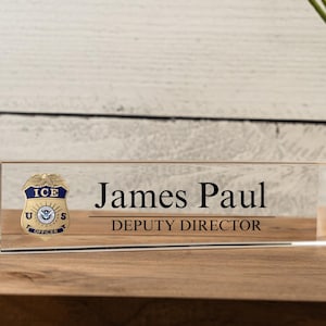 May include: A clear acrylic desk nameplate with a gold and blue ICE US Officer badge, the name "James Paul" and the title "Deputy Director".