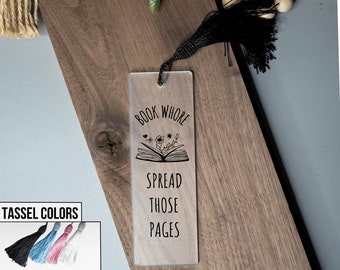 Op de afbeelding: Een transparante acryl boekenlegger met een zwarte kwast. De boekenlegger heeft de tekst "Book Whore Spread Those Pages" en een zwart-witte illustratie van een boek met bloemen.