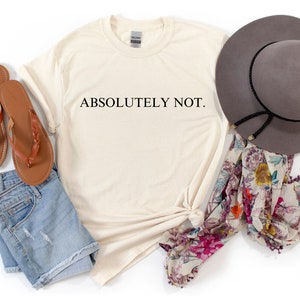 May include: A beige t-shirt with the text "ABSOLUTELY NOT." printed in black. The shirt is laid out on a white surface with a pair of brown sandals, a pair of denim shorts, a floral scarf, and a gray fedora.