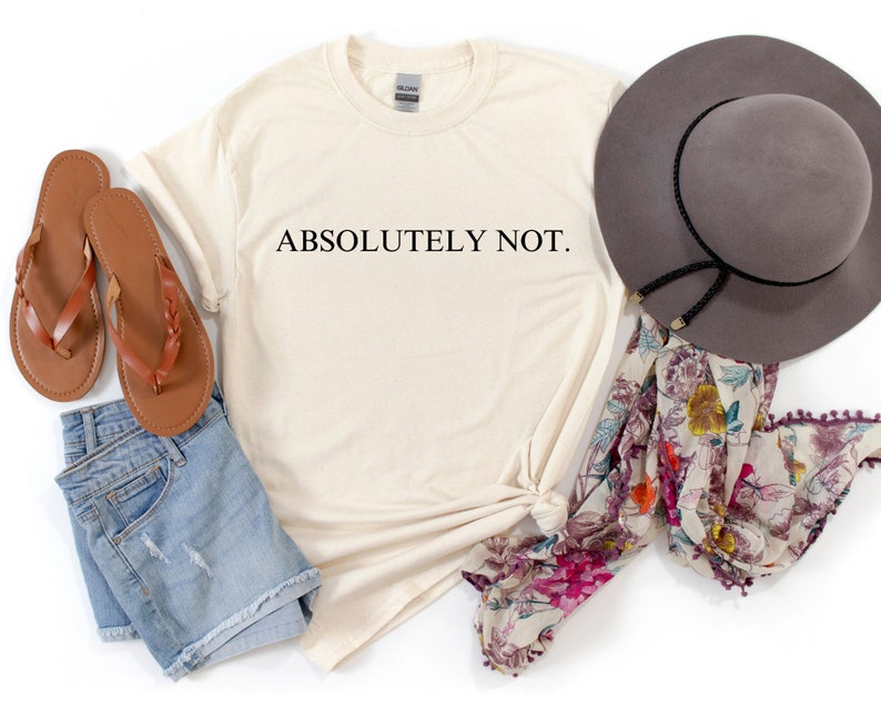 May include: A beige t-shirt with the text "ABSOLUTELY NOT." printed in black. The shirt is laid out on a white surface with a pair of brown sandals, a pair of denim shorts, a floral scarf, and a gray fedora.