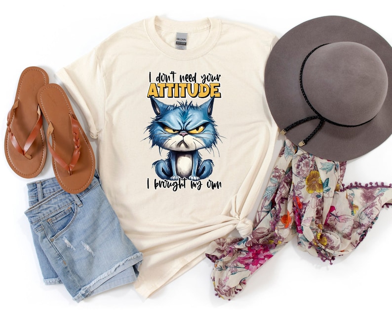Op de afbeelding: Een beige T-shirt met een blauwe kat cartoon afbeelding en de tekst "I don't need your attitude I brought my own". Het shirt ligt op een witte ondergrond met een bruine hoed, een paar bruine sandalen en een bloemen sjaal.