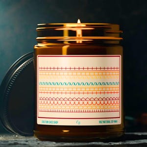 May include: A lit candle in a brown glass jar with a gold lid. The label features colorful geometric patterns and the text "CREATION SHED SHOP" and "9OZ NATURAL SOY WAX". The jar is partially open, revealing the metal lid.