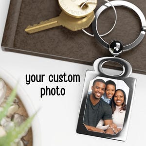 May include: Silver keychain with a photo frame featuring a family of three. The frame is rectangular with rounded corners and a small loop at the top. The photo shows a man, a woman, and a child smiling.