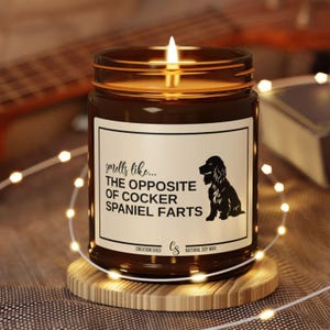 May include: A lit brown glass candle jar with a white label that reads "smells like... THE OPPOSITE OF COCKER SPANIEL FARTS" and a Cocker Spaniel silhouette. The candle sits on a wooden coaster, surrounded by fairy lights.