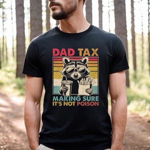 K&ouml;nnte beinhalten: Schwarzes T-Shirt mit einem Waschb&auml;r-Motiv, der eine Sonnenbrille tr&auml;gt und ein Getr&auml;nk und Pommes h&auml;lt. Der Text lautet "DAD TAX" und "MAKING SURE IT'S NOT POISON".