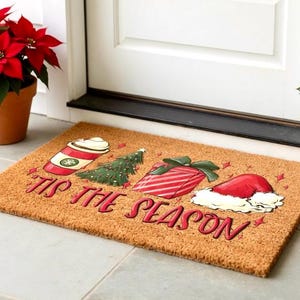 K&ouml;nnte beinhalten: Eine rechteckige Fu&szlig;matte mit festlichem Design. Sie zeigt einen Kaffeebecher, einen Weihnachtsbaum, ein verpacktes Geschenk und einen Weihnachtsm&uuml;tze. Der Text "'Tis the Season" ist in roter Schrift geschrieben. Die Matte besteht aus einem braunen, strukturierten Material.