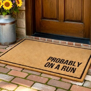 May include: A rectangular tan coir doormat with a thin black border and the black text "PROBABLY ON A RUN" printed in the lower right corner. The mat is made of natural fiber and features a coarse, textured surface for wiping footwear.