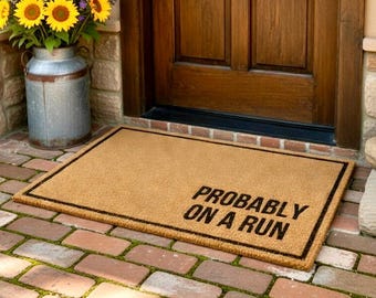 May include: A rectangular tan coir doormat with a thin black border and the black text "PROBABLY ON A RUN" printed in the lower right corner. The mat is made of natural fiber and features a coarse, textured surface for wiping footwear.