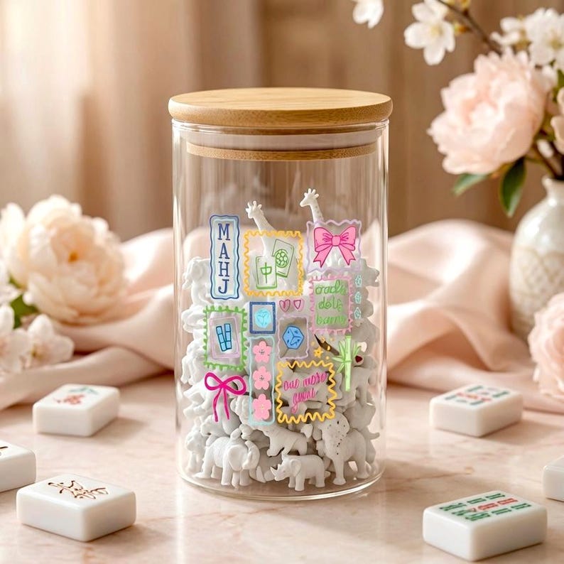 May include: A clear glass cylindrical storage jar with a light wood lid, filled with small white animal figurines. The exterior of the glass features colourful, whimsical sticker-style illustrations of bows, flowers, and Mahjong-related symbols.