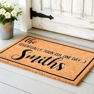May include: A rectangular coir doormat with a black border and the family name "Smiths" in elegant black script. The text "the (eventually, soon-ish, one day...)" is above the name. The mat is on a white wooden porch.