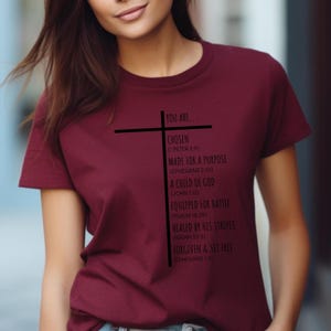 May include: A maroon t-shirt with a black cross and text that reads "You are... Chosen (1 Peter 2:9) Made for a Purpose (Ephesians 2:10) A Child of God (John 1:12) Equipped for Battle (Psalm 18:39) Healed by His Stripes (Isaiah 53:5) Forgiven & Set Free (Ephesians 1:7)"