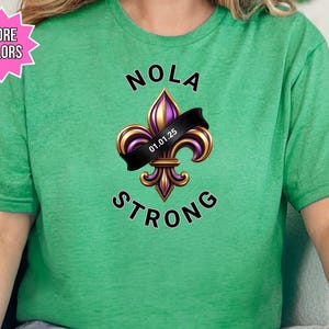 May include: Green t-shirt with the words "NOLA STRONG" and a fleur-de-lis design in gold, purple, and black. The fleur-de-lis has a black banner with the date "01.01.25". A pink starburst graphic says "MORE COLORS".