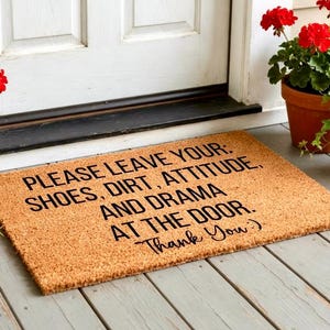 May include: A rectangular coir doormat with the message "PLEASE LEAVE YOUR: SHOES, DIRT, ATTITUDE, AND DRAMA AT THE DOOR. Thank You:)" in black text. The mat is placed in front of a white door, with red flowers in terracotta pots.