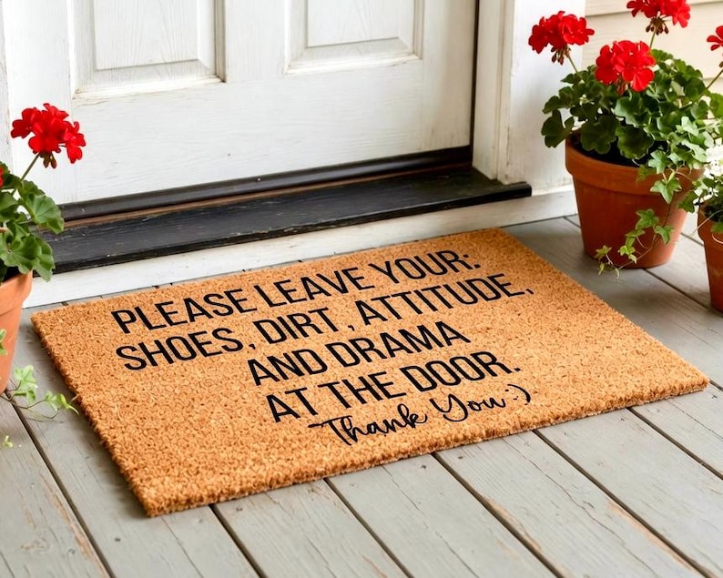 May include: A rectangular coir doormat with the message "PLEASE LEAVE YOUR: SHOES, DIRT, ATTITUDE, AND DRAMA AT THE DOOR. Thank You:)" in black text. The mat is placed in front of a white door, with red flowers in terracotta pots.