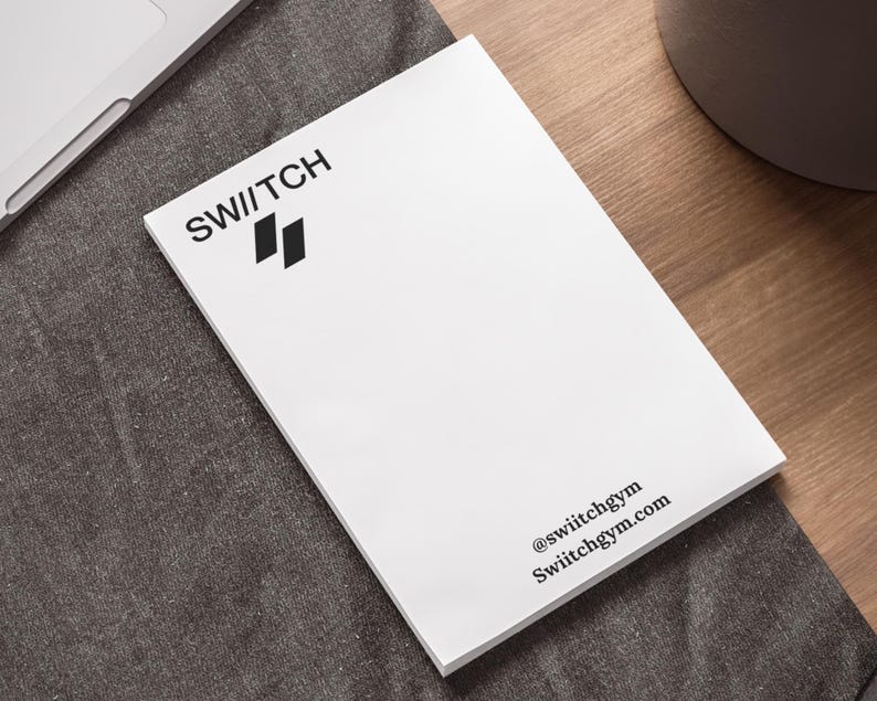 以下が含まれることがあります： 「SWITCH」というテキストと二重線のロゴが書かれた白いメモ帳。メモ帳には「@switchgym」と「Switchgym.com」のテキストも含まれています。