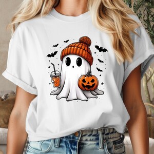 May include: White t-shirt featuring a cartoon ghost wearing an orange and white knitted hat, holding a pumpkin and a cup of orange juice. The ghost is surrounded by small black bats and stars.