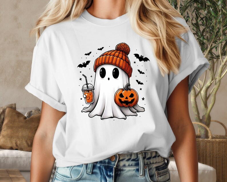 May include: White t-shirt featuring a cartoon ghost wearing an orange and white knitted hat, holding a pumpkin and a cup of orange juice. The ghost is surrounded by small black bats and stars.
