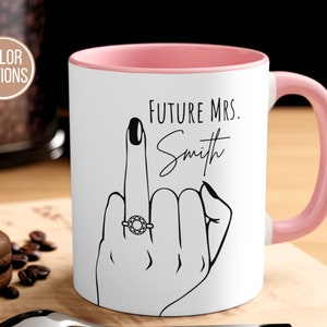 May include: White ceramic mug with a pink rim and handle. The mug features a black and white illustration of a hand with a ring on the middle finger, and the text "FUTURE MRS. Smith".