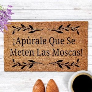 May include: A rectangular doormat made of natural coir fiber, featuring the phrase "¡Apúrale Que Se Meten Las Moscas!" in black lettering. The mat is bordered with black leaf designs and rests on a white wooden surface. A cup of coffee and shoes are in the image.