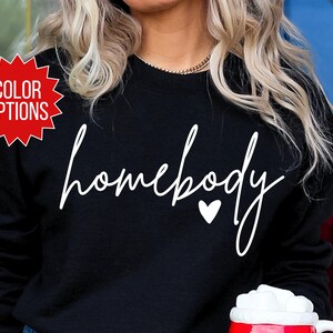 K&ouml;nnte beinhalten: Schwarzer Pullover mit wei&szlig;em Text, der "homebody" mit einem kleinen wei&szlig;en Herz sagt.