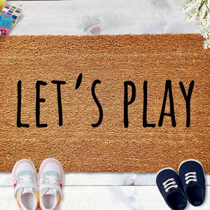 May include: A brown doormat with the text "LET'S PLAY" in black lettering. Two pairs of shoes are on either side of the doormat, one pair is white and blue sneakers and the other pair is navy blue sneakers.