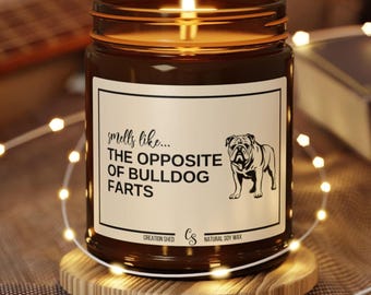 May include: A lit brown glass jar candle with a white label featuring the text "smells like... THE OPPOSITE OF BULLDOG FARTS" and a bulldog illustration. The candle sits on a wooden coaster, surrounded by string lights.