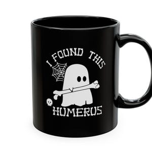 Op de afbeelding: Een zwarte keramische mok met een witte afbeelding van een geest die een bot in zijn mond houdt. Het ontwerp bevat een spinnenweb, een kleine schedel en de tekst "I FOUND THIS HUMERUS" in een gestileerd lettertype dat op botten lijkt.