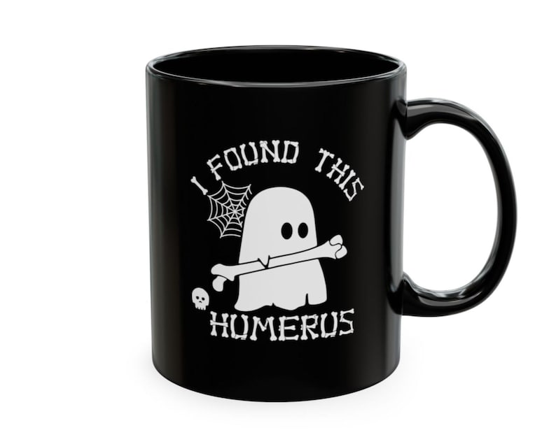 Op de afbeelding: Een zwarte keramische mok met een witte afbeelding van een geest die een bot in zijn mond houdt. Het ontwerp bevat een spinnenweb, een kleine schedel en de tekst "I FOUND THIS HUMERUS" in een gestileerd lettertype dat op botten lijkt.