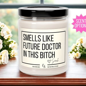 May include: A white candle with a black label that reads "Smells Like Future Doctor In This Bitch" with a heart and the name "Sarah" written below. The candle is 9 oz and made with natural soy wax.