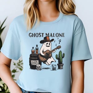 May include: A light blue t-shirt with a graphic of a ghost wearing a cowboy hat and playing a guitar. The text "Ghost Malone" is printed above the ghost.