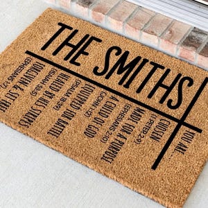 May include: A rectangular coir doormat with the text "THE SMITHS" in large, black, stylized letters. Below, a grid lists inspirational phrases and Bible verses, including "Chosen, You Are." The mat is a natural brown color.