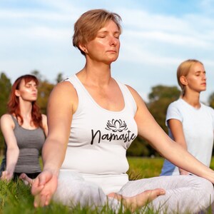 May include: Three people are sitting in a field of green grass with their eyes closed, meditating. The person in the foreground is wearing a white tank top with the word "Namaste" and a lotus flower design. The other two people are wearing white shirts.
