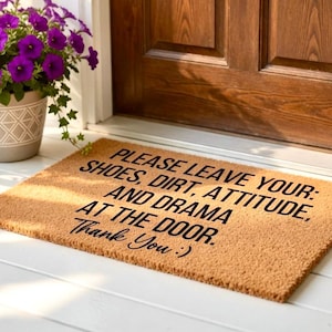 May include: A rectangular doormat made of natural fibres, with the text "PLEASE LEAVE YOUR: SHOES, DIRT, ATTITUDE, AND DRAMA AT THE DOOR. Thank You :)" in black. The mat is placed in front of a wooden door.