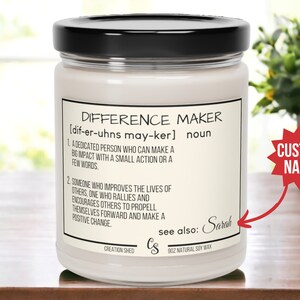 May include: A white candle jar with a black lid and a label that reads "Difference Maker" with a definition and the word "Sarah" written in cursive. The label also includes the text "Creation Shed" and "9oz Natural Soy Wax". A red starburst with the text "Custom Name" is in the background.
