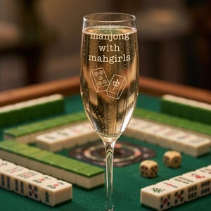 K&ouml;nnte beinhalten: Ein Sektglas aus klarem Glas, gef&uuml;llt mit einem sprudelnden Getr&auml;nk. Das Glas ist mit dem wei&szlig;en Schriftzug "manjong with mahgirls" und einer grafischen Darstellung von zwei Mahjong-Steinen versehen, von denen einer Punkte zeigt.