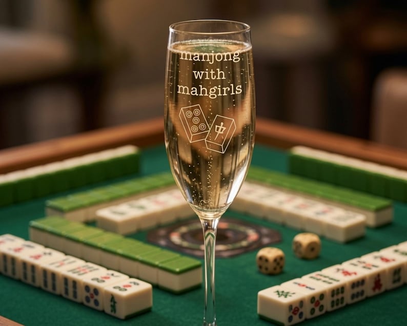 K&ouml;nnte beinhalten: Ein Sektglas aus klarem Glas, gef&uuml;llt mit einem sprudelnden Getr&auml;nk. Das Glas ist mit dem wei&szlig;en Schriftzug "manjong with mahgirls" und einer grafischen Darstellung von zwei Mahjong-Steinen versehen, von denen einer Punkte zeigt.