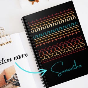 May include: Black spiral-bound notebook with a colorful geometric design on the cover. The design includes orange, yellow, teal, and red lines. The name "Samantha" is written in teal script. The notebook is open to a page with the words "custom name".