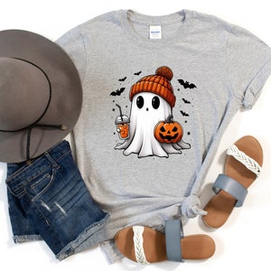 May include: A grey t-shirt featuring a cartoon ghost wearing a pumpkin-coloured beanie and holding a pumpkin and a cup of coffee. The ghost has large eyes and a friendly expression. The shirt is decorated with bats and stars.