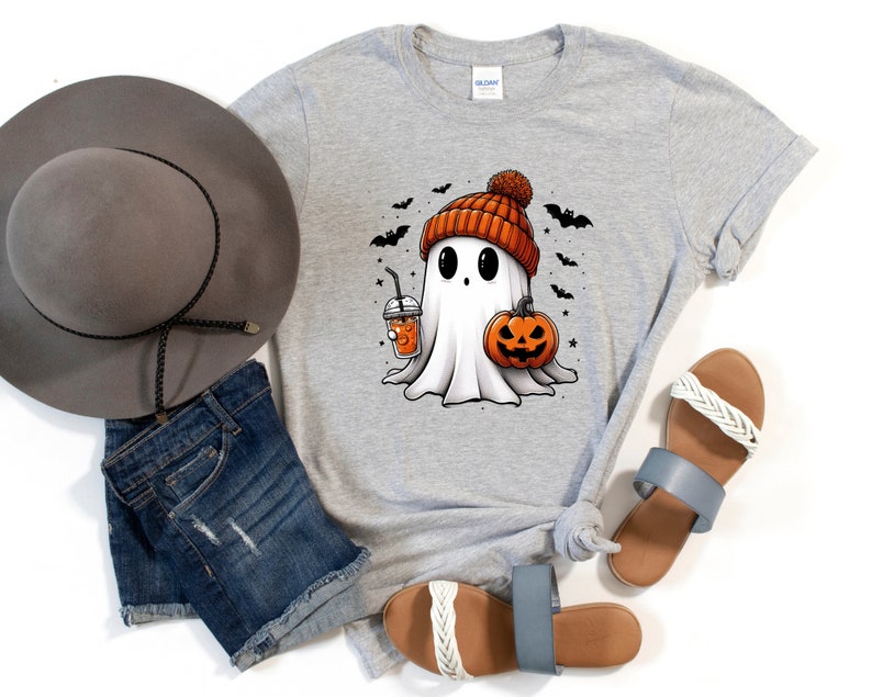 May include: A grey t-shirt featuring a cartoon ghost wearing a pumpkin-coloured beanie and holding a pumpkin and a cup of coffee. The ghost has large eyes and a friendly expression. The shirt is decorated with bats and stars.