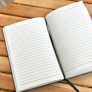 May include: An open, lined notebook with a black cover and a black ribbon bookmark. The pages are white with horizontal black lines. The notebook is on a wooden surface.