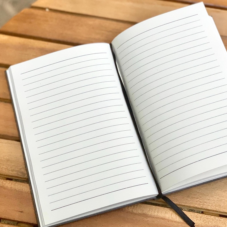 May include: An open, lined notebook with a black cover and a black ribbon bookmark. The pages are white with horizontal black lines. The notebook is on a wooden surface.
